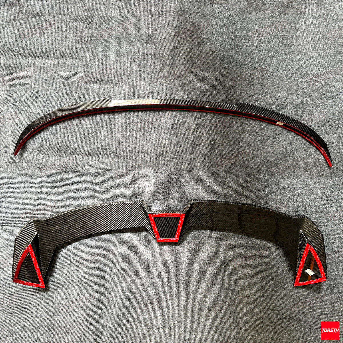 A pair of carbon fiber rear spoiler winglets for a vehicle, displayed flat on a textured gray background. The top piece is a simple curved winglet, while the bottom piece is a more complex multi-piece winglet set with red stitched trim accents.