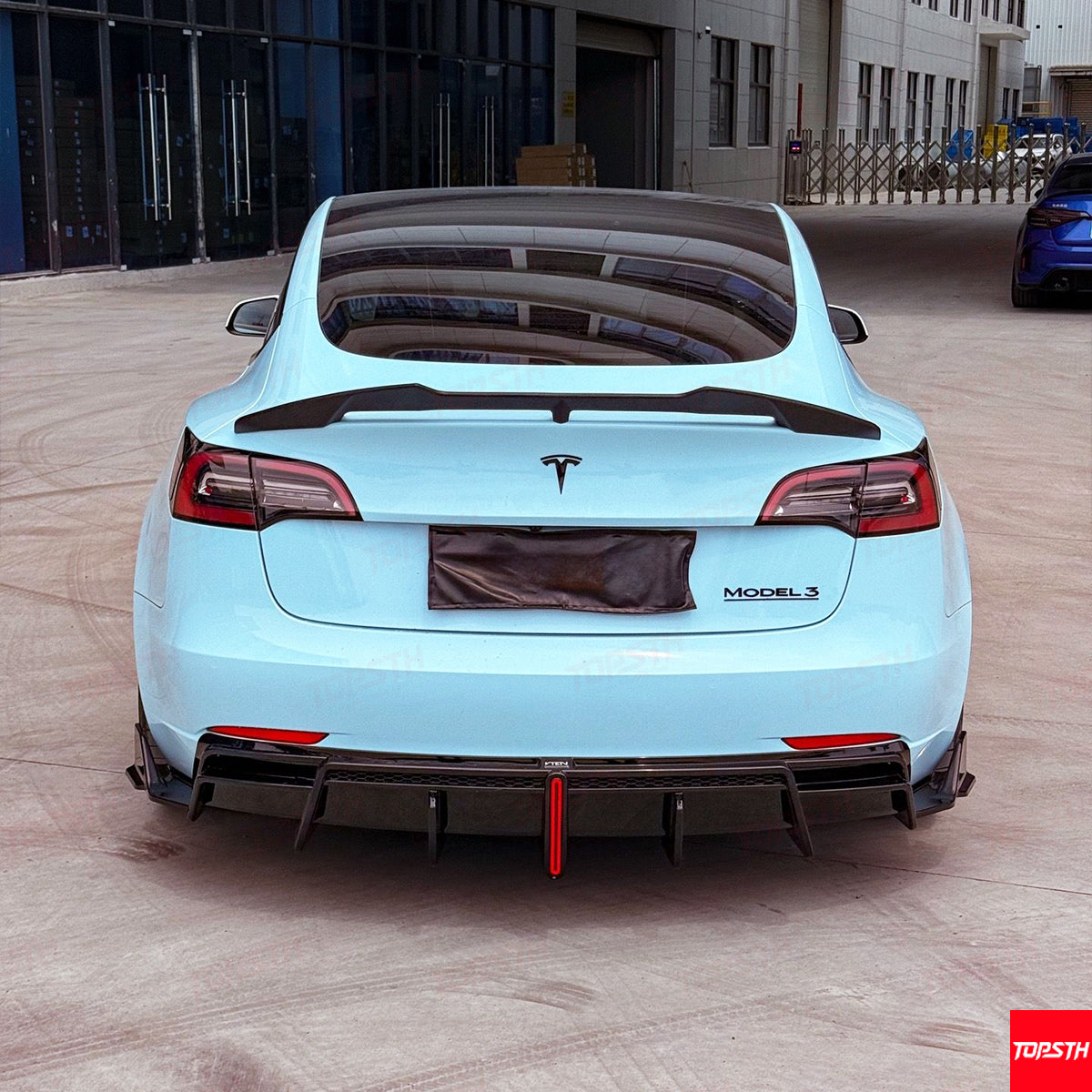 Back view of a light blue Tesla Model 3 in an industrial setting