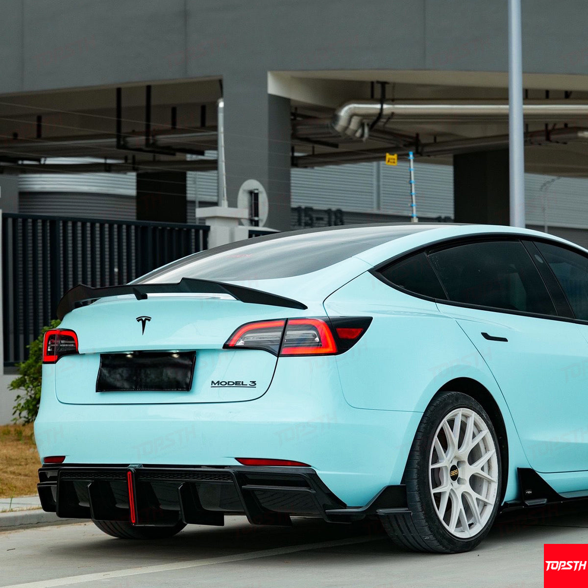 Light blue Tesla Model 3 parked in an outdoor setting with a building in the background.